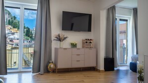 A spacious living room with a large TV mounted on the wall, a wooden cabinet with drawers, and a view of a hillside through large sliding glass doors.