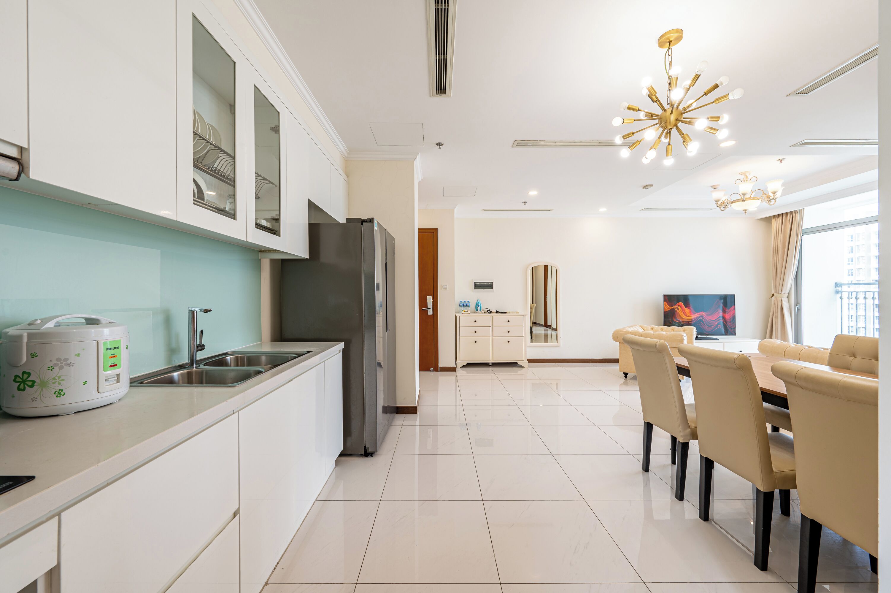 A modern kitchen with a built-in refrigerator, a sink, and a countertop with a rice cooker. There is a dining area with a table and chairs, and a large window with curtains.