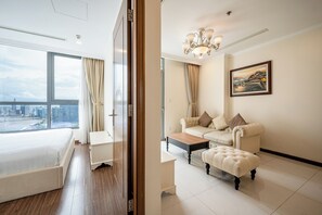 A modern hotel room with a large bed, a seating area with a sofa and ottoman, a dining table, and a city view through large windows.