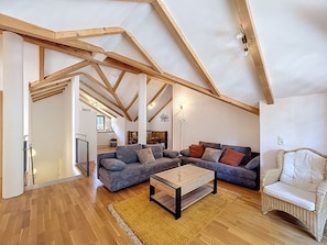 Un loft spacieux avec des poutres apparentes, un grand canapé d’angle gris, une petite table basse en bois et un fauteuil en osier.