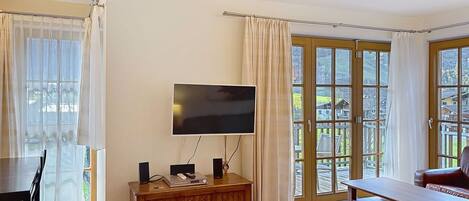 A living room with a TV, wooden furniture, a dining table, and large windows with curtains.