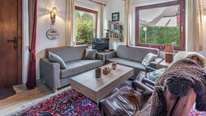 A cozy living room with a grey sofa, a wooden coffee table, and a leather armchair. There is a bookshelf with books and a television on a stand. Large windows offer a view of greenery outside.