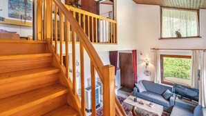 A spacious living area with a wooden staircase, a sofa, a television, and a view of greenery outside.