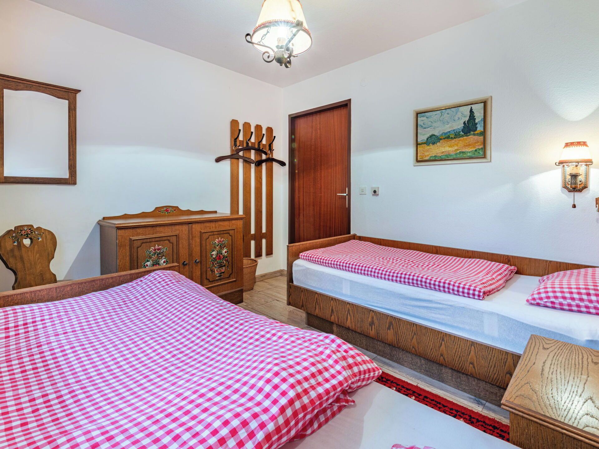 A room with two beds, a wooden wardrobe, a door, a painting, and a wall-mounted lamp.