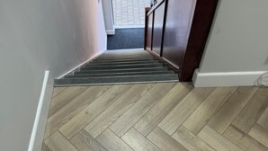 Un suelo de madera con un diseño de espiga guía hacia una escalera con barandilla de madera oscura y paredes blancas.