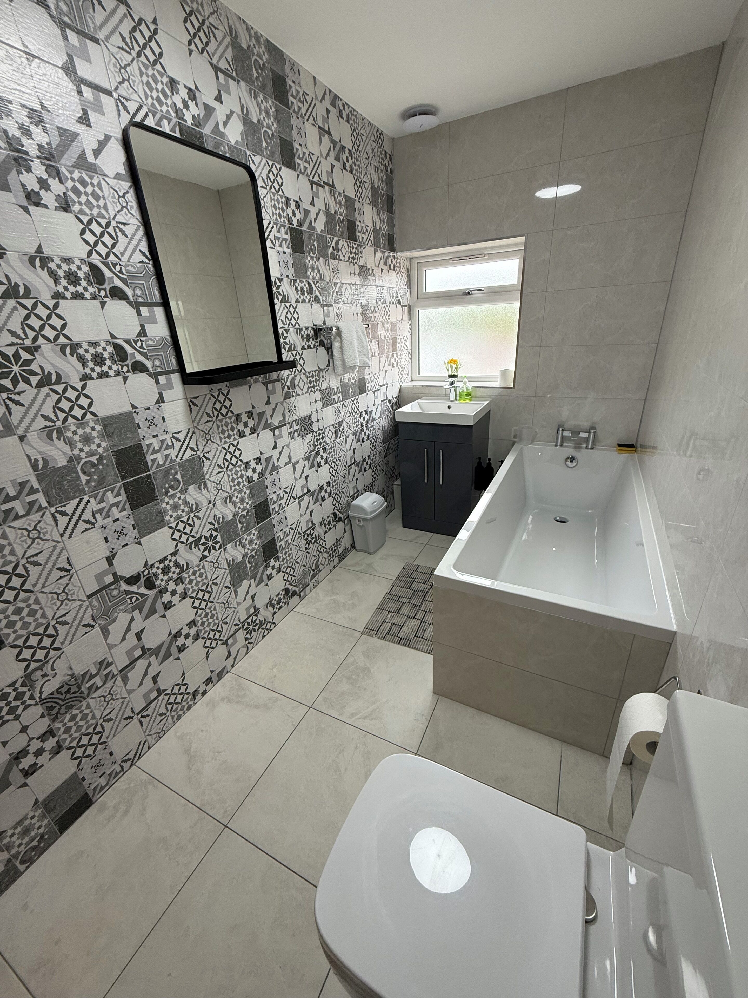 Ein modernes Badezimmer mit gemusterten Fliesen, einer großen Badewanne, einer Toilette und einem Waschbecken mit Fenster darüber.