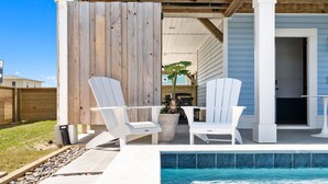 A covered outdoor area with a pool, white Adirondack chairs, and a wooden fence.