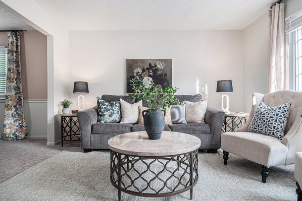 A living room with a grey sofa, a round coffee table, and a beige armchair. There are two side tables with lamps, a large painting on the wall, and a window with curtains.