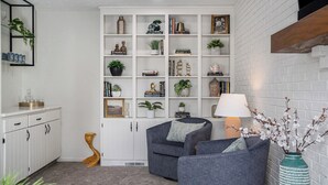 A cozy living room with a grey sofa, a white bookshelf, and a decorative vase with branches. - Warm & Spacious 4BR Retreat w/ Hot Tub & Grill! (Omaha)