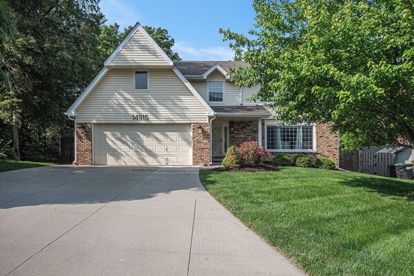 A two-story house with a garage, brick facade, and a well-maintained lawn. - Warm & Spacious 4BR Retreat w/ Hot Tub & Grill! (Omaha)