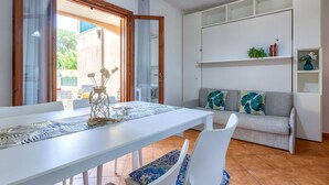Un coin repas avec une table et des chaises blanches, un canapé agrémenté de coussins à motifs et une étagère blanche où sont disposés des objets décoratifs.