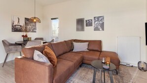 Een moderne woonkamer met een bruine hoekbank, een ronde salontafel en een eethoek met een kleine tafel en stoelen.