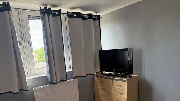 A room with a television on a wooden cabinet, a window with curtains, and a radiator. - 6 Guests - 3 Bedrooms - Close to London Center (London)