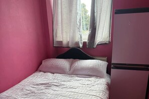 Um quarto pequeno com uma cama de casal, paredes cor-de-rosa e uma janela com cortinas.