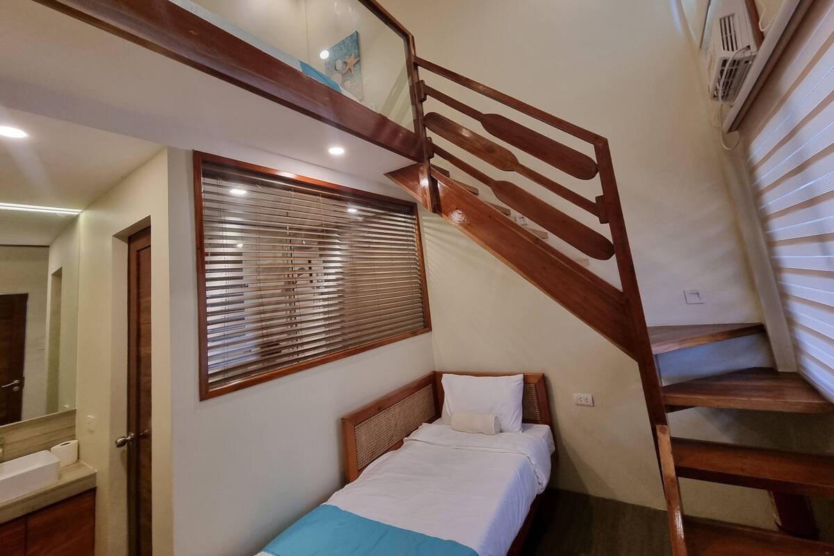 Un dormitorio con cama de madera, ventana con persianas y una escalera de madera, con un baño visible al fondo.