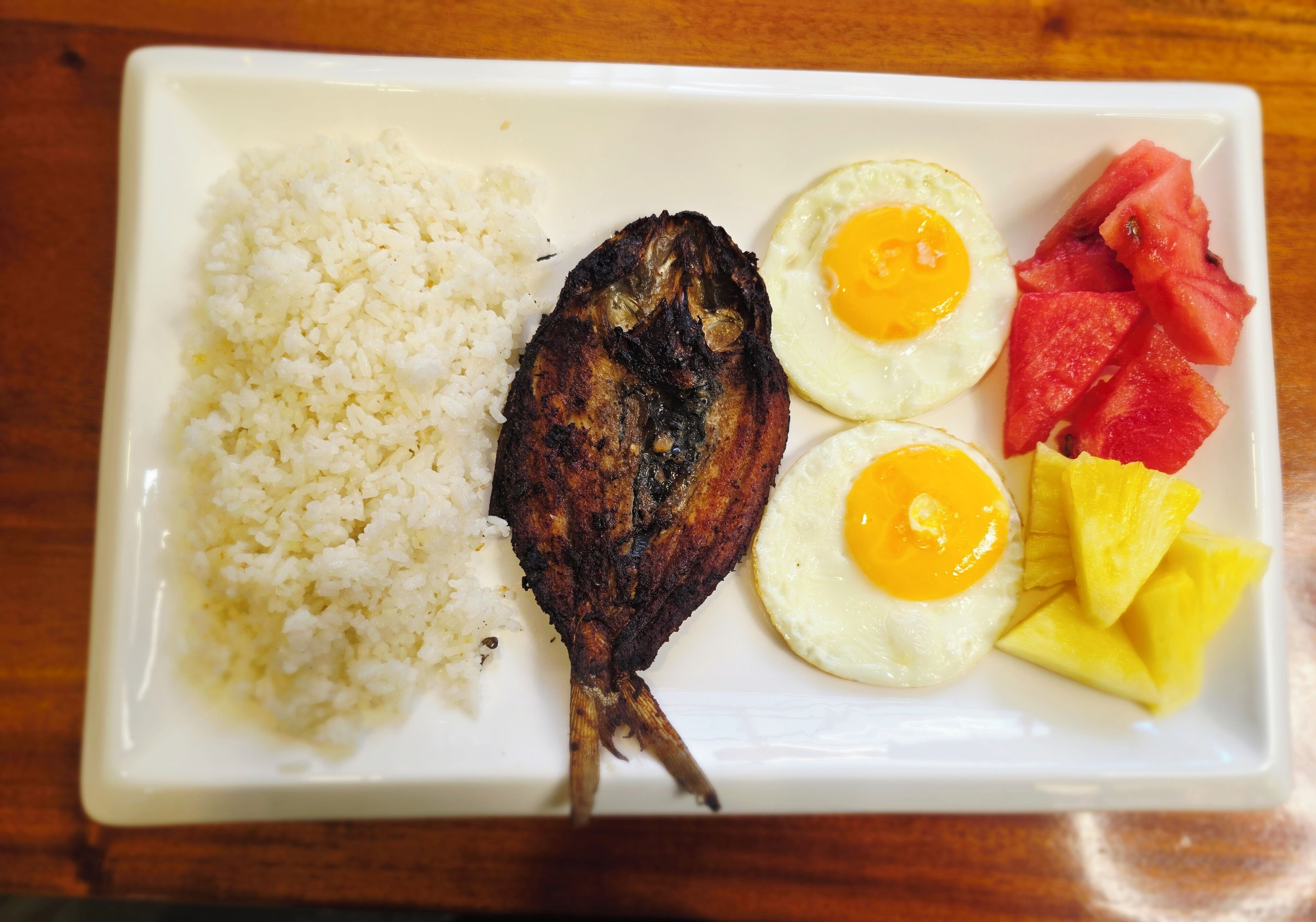 A plate with white rice, two fried eggs, a grilled fish, and watermelon and pineapple slices.