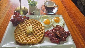 A breakfast plate with waffles, eggs, bacon, and grapes on a table with a lamp, a small plant, and a cup of coffee. - The CLiffhouse Samboan (Samboan)
