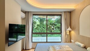 A modern bedroom with a large bed, a flat-screen TV, and a view of greenery outside.