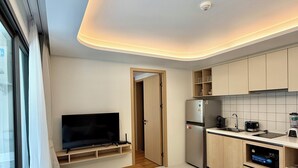 A modern living room with a sofa, a flat-screen TV, and a wooden coffee table. Adjacent is a kitchen with a refrigerator, stove, and microwave.