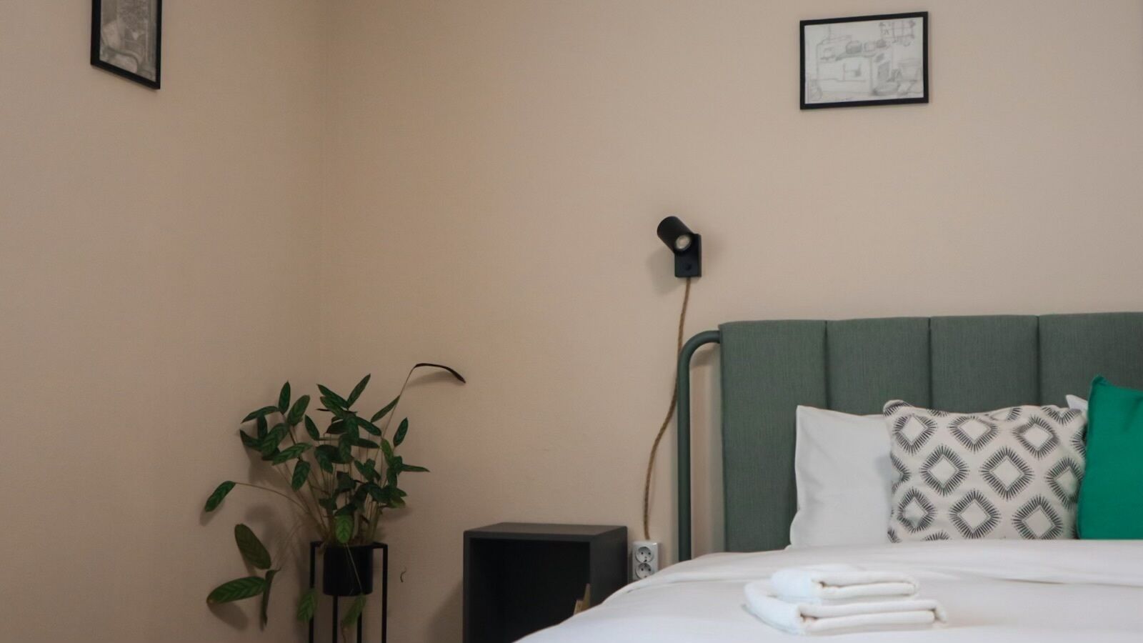 A neatly made bed with a patterned pillow, a bedside plant, and a speaker on a bedside table in a hotel room.