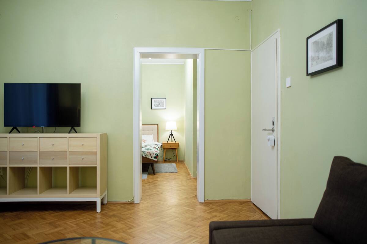 A room with a TV on a stand, a bed with a nightstand, a door, and a framed picture on the wall.