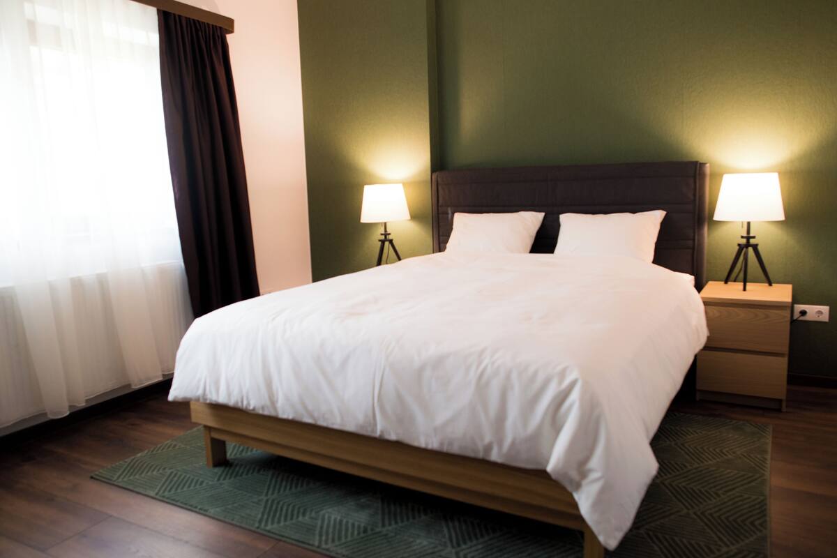 A neatly made bed with white linens, two bedside tables with lamps, and a green patterned rug.