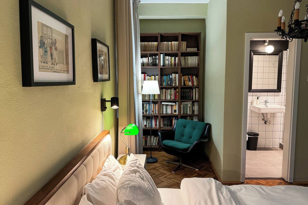 A hotel room with a bed, a green chair, a bookshelf, and a bathroom visible through an open door.