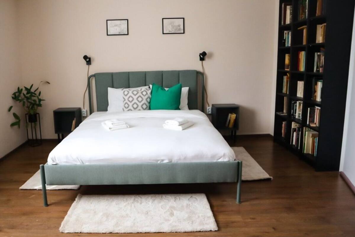 A neatly arranged bedroom with a bed, bedside tables, a bookshelf, and a potted plant.