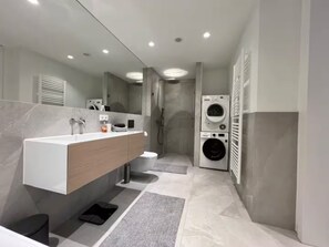 A modern bathroom with a large mirror, a sink with a wooden vanity, a toilet, a shower, and a stacked washer and dryer. - Ziegelhofring Apartment (Bad Vilbel)
