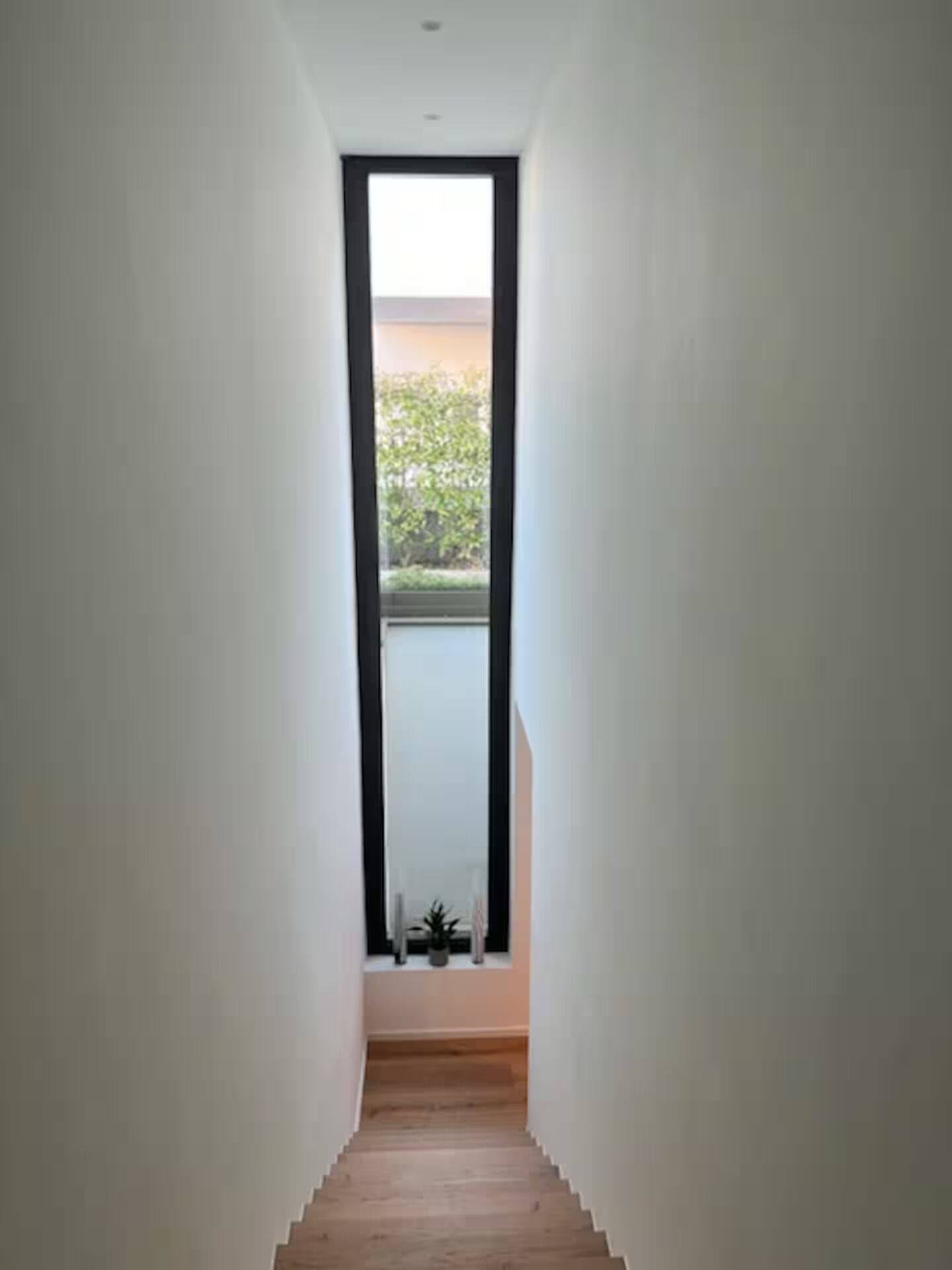 Een smalle gang met houten vloer, witte muren en een groot raam aan het einde.