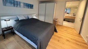 A modern bedroom with a large bed, a bedside table, a wardrobe, and a bathroom visible through an open door. - Apartment Ziegelhofring (Bad Vilbel)