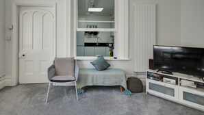 Een kamer met een witte deur, een raam met uitzicht op een keuken, een televisie op een witte standaard en een grijze stoel met kussen.