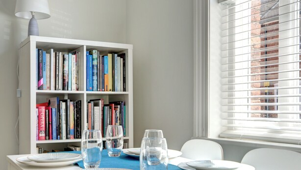Een eetruimte met een witte tafel gedekt voor vier personen, een boekenkast op de achtergrond en een raam met jaloezieën.