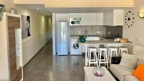 Een moderne keuken met witte kastjes, een ingebouwde oven en een spoelbak. Er is een eethoek met een witte tafel en barkrukken.
