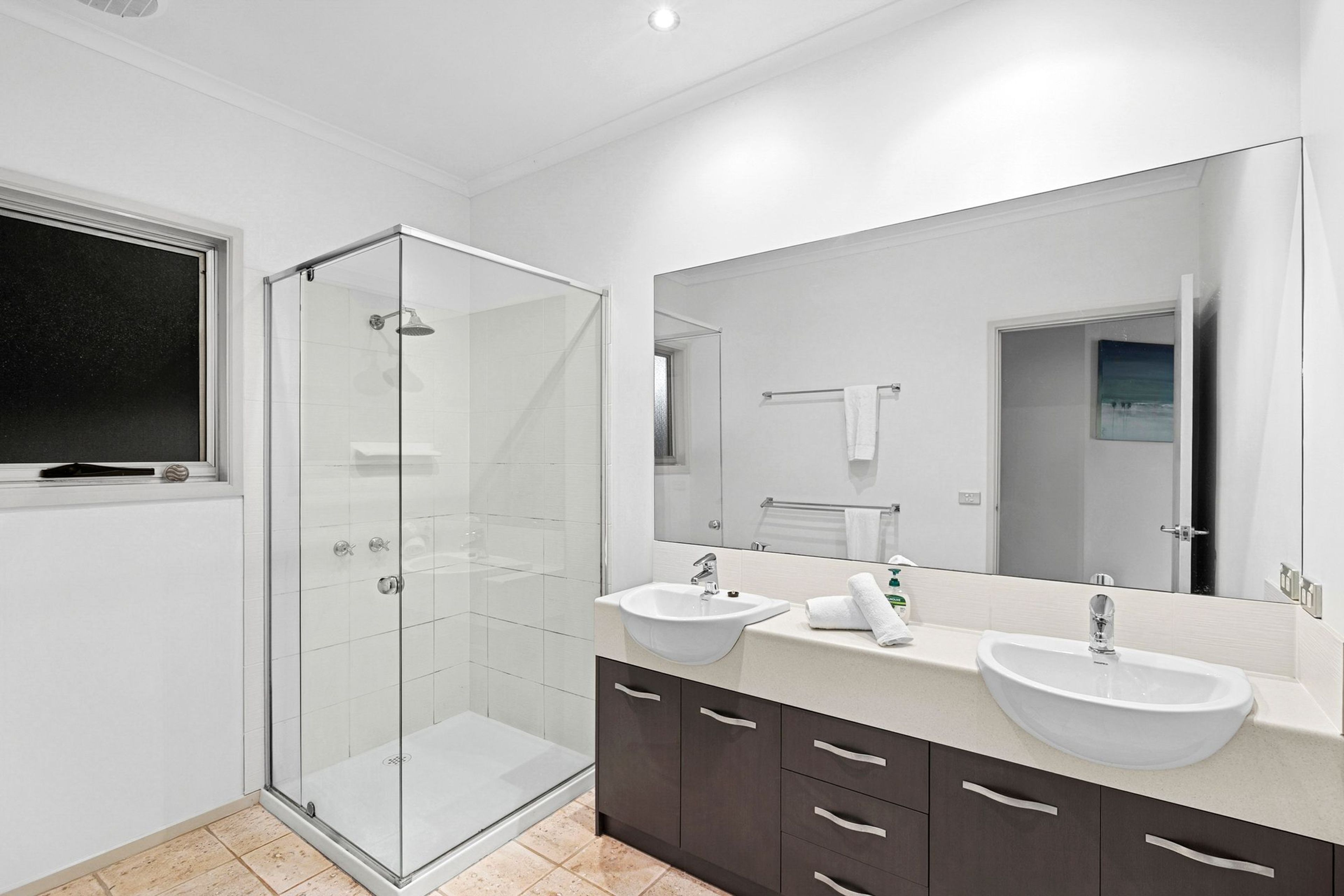 A modern bathroom with a glass-enclosed shower, two sinks, a large mirror, and dark cabinetry.
