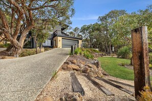 A modern house with a paved driveway leading to a garage, surrounded by mature trees and a well-maintained lawn.