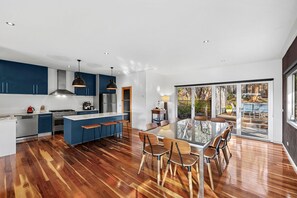 A modern kitchen with a central island, wooden flooring, and a dining area with a large table and chairs. - Orchid Haven (Fairhaven)