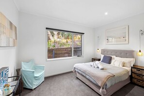 A bedroom with a large bed, a window with a view of greenery, a bedside table with a lamp, and a wall-mounted artwork.