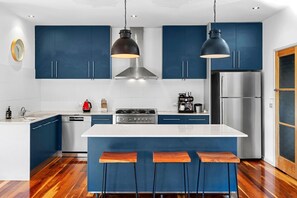 A modern kitchen with blue cabinets, a stainless steel refrigerator, a gas stove, and a bar area with stools.