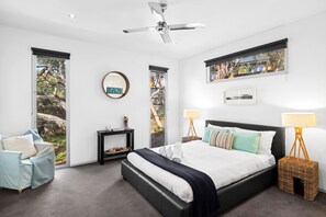 Une chambre moderne avec un grand lit, deux tables de chevet, un ventilateur de plafond et de grandes fenêtres donnant sur des arbres et un immeuble.