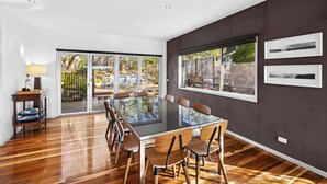 A modern dining area with a glass table and wooden chairs, large windows with a view of the outdoors, and a dark wall with two framed pictures. - Orchid Haven (Fairhaven)