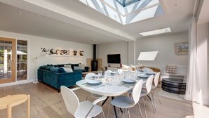 Una sala de estar moderna con techo alto y tragaluces, un comedor con mesa redonda y sillas, y una zona de estar acogedora con un sofá azul.