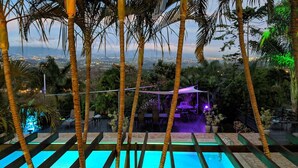Zona de piscina al aire libre con vista a un paisaje urbano al anochecer, rodeada de palmeras y un patio con mobiliario.