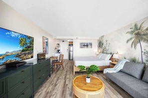 A modern hotel room with a bed, a sofa, a dining area, a TV, and a painting of a beach scene. - Waikiki studio w/ lanai and beach access (Honolulu)