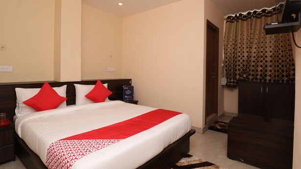 A neatly made bed with red pillows, a wooden headboard, and a red and white patterned bedspread. There is a dark wooden wardrobe to the right and a small table with a lamp to the left. - HOTEL O MDA INN (Lucknow)