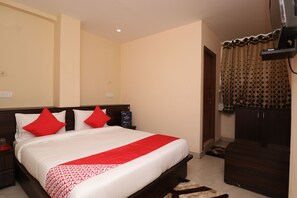 Una cama bien hecha con almohadones rojos, un cabecero de madera y una colcha con estampado rojo y blanco. A la derecha se ve un armario de madera oscura y, a la izquierda, una mesita de noche con una lámpara.