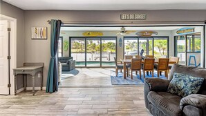 A spacious living area with a view of a pool and outdoor area, a dining set, and a sign reading "LET'S DO SUNSET".
