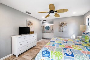 Un dormitorio con una cama, un televisor sobre un aparador, un ventilador de techo y un baño con lavabo y espejo.