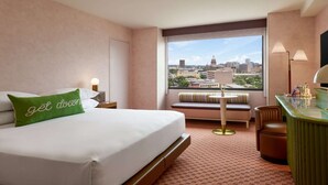 Hotellihuone, jossa on sänky, penkki, työpöytä ja suuri ikkuna, josta avautuu näkymä kaupunkiin.