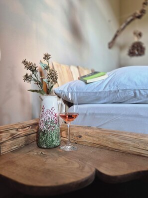 Una mesa de madera con un jarrón de flores secas y un vaso de bebida rosa, sobre una cama con un cojín azul y un libro.
